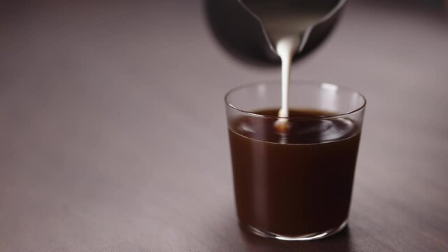 Slow motion milk pour into tumbler glass with coffee on walnut table