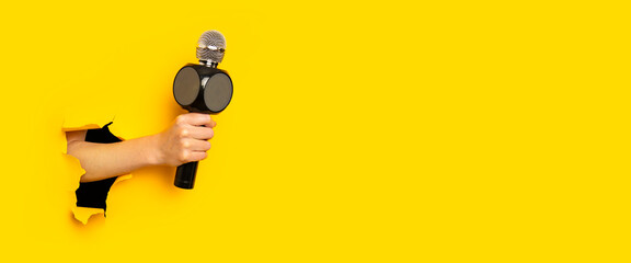 Female hand holding a microphone in a hole in a torn bright yellow cardboard background. Banner