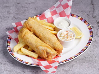 Classic fish and chips with crispy battered fish fillets, golden fries, coleslaw, tartar sauce, and a lemon wedge, served on a checkered paper-lined plate.