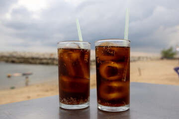 glass of iced cola sits on a table by the sea, with a cloudy sky backdrop. The drink's bubbles and condensation are visible