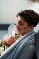 Sick man wrapped in a blanket is doing inhalation by nebulizer for the cough treatment. Patient is...