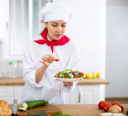 Cheerful female chef in white uniform preparing vegetable dish in private kitchen