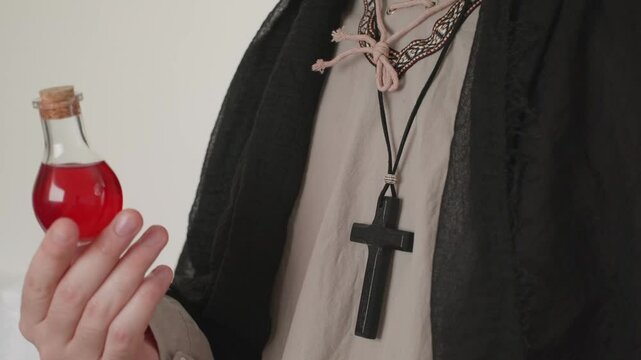 A man with a cross around his neck holding a potion in a red glass bottle