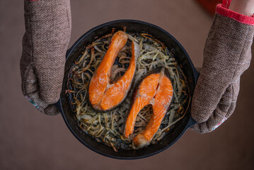 Chef, wearing oven mitts, holds a cast iron skillet containing two roasted salmon steaks with...