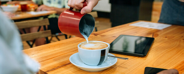 Banner of unrecognizable barista preparing a frothy cappuccino, skillfully pouring creamy milk into a coffee cup on a wooden table in a cozy cafe, while a customer waits nearby