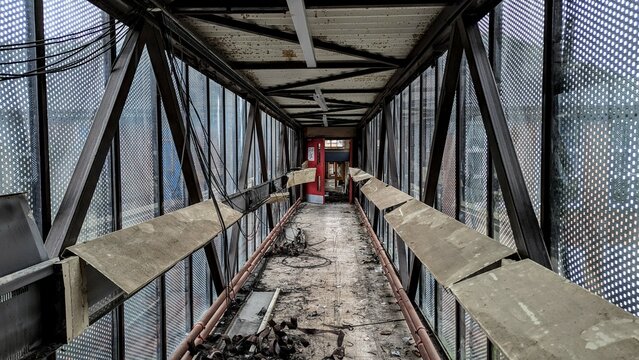 Collapsing eerie footbridge structure falling apart. Wrecked hallway corridor, twisted rusting metal. Broken windows disaster inside creepy walkway tunnel. Abandoned empty passageway gloomy skybridge.