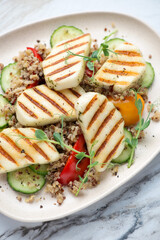 Slices of grilled halloumi cheese with quinoa, bell pepper and cucumber on a beige plate, vertical shot, middle close-up