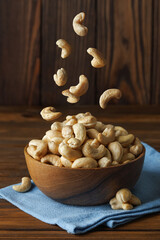 Cashew nuts falling into bowl placed on blue napkin on rustic wooden table, creating an enticing arrangement of beloved snack rich in protein, vitamins and minerals