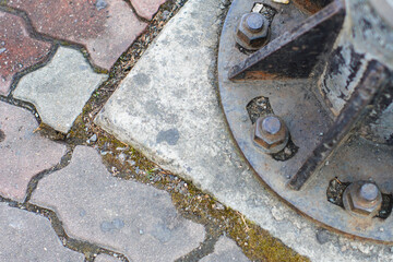 A close-up of a rusty metal base with bolts on a weathered concrete and brick pavement, showing...