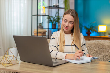 Caucasian woman sits at home on table typing on laptop, smiling, remote education, notebook open,...
