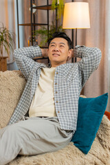 Happy carefree middle-aged Chinese man sitting comfortably with hand behind head on sofa in living...