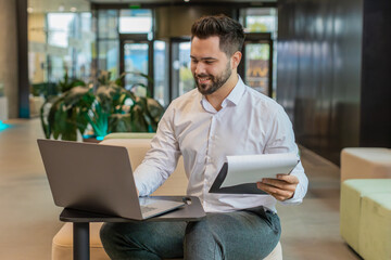 Caucasian young businessman using laptop and working with documents, analyzing information, reading papers prepare financial report. Male entrepreneur in formal analyzing graph in modern office lobby.