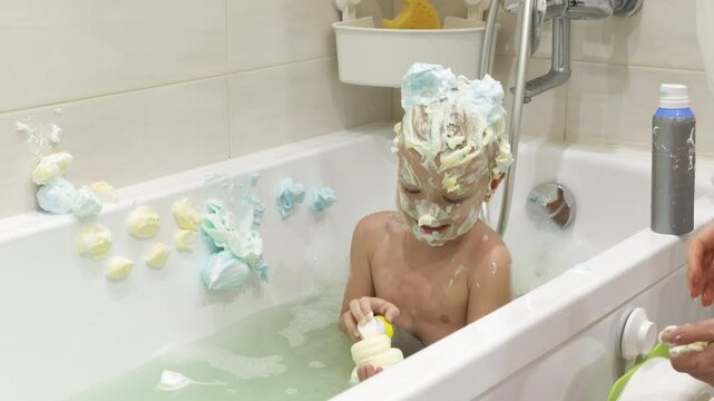 young boy has fun in the bathtub with whipped cream playing with the creamy foam and looks very happy.