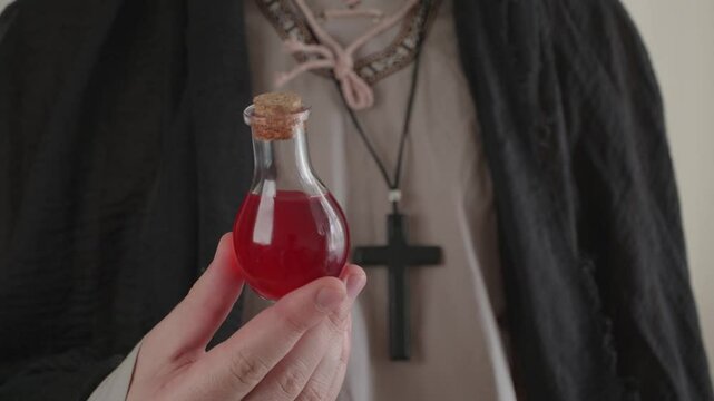A man with a cross around his neck holding a potion in a red glass bottle