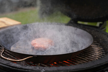 Fresh meat cutlet in a frying pan grill. A large round wood-burning grill is roasting beef patty for burger. Barbecue festival in the city park. Street fast food.