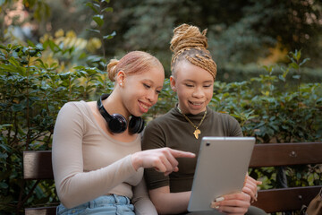 Two smiling albino African female students sitting together on a park bench, using a tablet. One...