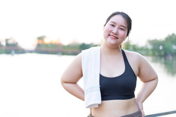 Fototapeta premium Cheerful attractive young overweight woman weight loss concept, asian smiling woman in sports bra, enjoying outdoors. She holds towel over her shoulder, radiating confidence and positivity