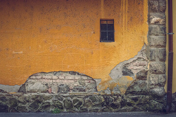 An old residential building wall in Szentendre, Hungary, with peeling plaster revealing brick and stone layers. A small window adds character. The textured facade shows age, history, and urban decay