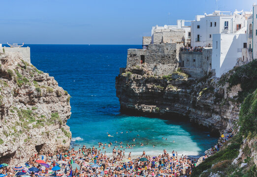 Crowded Lama Monachile beach with sunbathing vacationers, crystal waters, rocky cliffs, white buildings framing scenic coastal landscape in Polignano a Mare, southern Italy