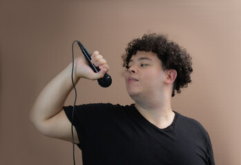 Young man with curly hair holds a microphone, looking to the side. Wearing a black t-shirt, standing against a neutral background. Confident expression, singer or performer concept, music, vocal.