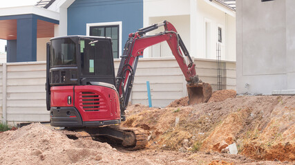 A small red excavator is digging a hole to prepare for building a wall around a concrete house on background Concept of working with machinery at a construction site.