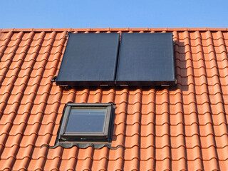 close-up of a red tiled roof featuring two black solar panels and a small skylight window under a clear blue sky. The image highlights sustainable energy solutions in modern housing