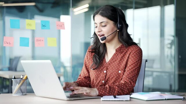 Young smiling female corporate operator in wireless headset working in customer support service on helpline telesale talking using laptop in business office. Agent of call center advises client online
