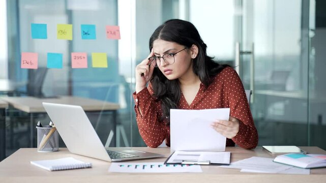 Confused young businesswoman having difficulty with paper work checking documents on laptop computer sitting at workplace in business office. Frustrated female financier unhappy with financial results