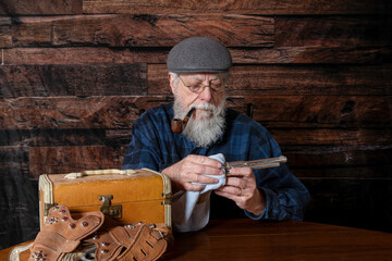 old man with white beard smoking pipe sitting at table cleaning vintage toy cap gun with dark wood background