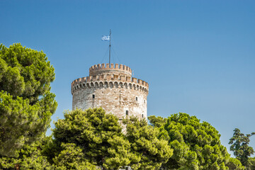 White tower of Thessaloniki, the Ottoman fortress, now an interactive exhibition of the city's history. Upper part of the tower with greek flag against blue clear sky and behind green pines.