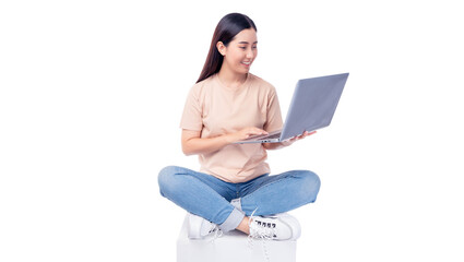 Cheerful young asian woman sitting cross-legged on a white cube while using a laptop. She wears T-shirt and blue jeans, smiling as she interacts with the mobile phone isolated on white background