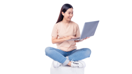 Cheerful young asian woman sitting cross-legged on a white cube while using a laptop. She wears T-shirt and blue jeans, smiling as she interacts with the mobile phone isolated on white background