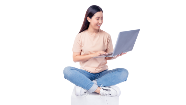 Cheerful young asian woman sitting cross-legged on a white cube while using a laptop. She wears T-shirt and blue jeans, smiling as she interacts with the mobile phone isolated on white background