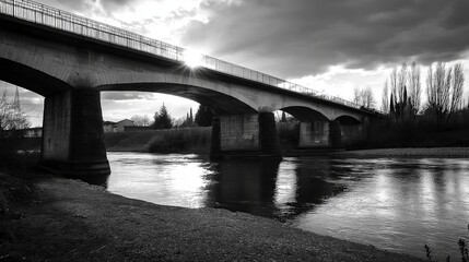 A black and white image of a bridge spanning a river, with dramatic shadows