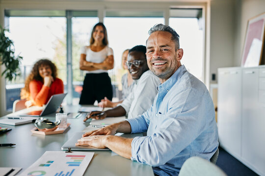 A group of smiling diverse business people are sitting in a meeting, in a conference room. They all look happy and smiling into the camera. On the table are graphs and a laptop.