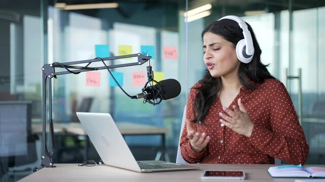 Young businesswoman with a microphone and headphones is recording a podcast using laptop sitting in business office. Female coach conducts remote training. Vlogger communicates online via video call