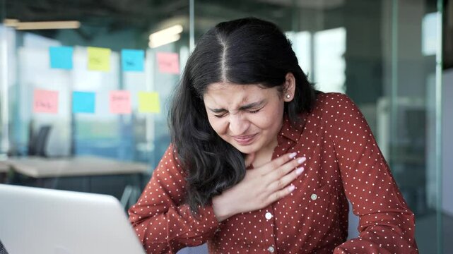 Young businesswoman has a heart attack while sitting at workplace in a business office. Sick female employee suffers from tension in the chest and massages the sore spot with her hand. Close up