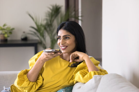 Indian woman relaxing on sofa holds smartphone leaves audio message, sending voicemail to friend, lead remote talk through speakerphone, using virtual assistant. Digital communication, modern AI tech