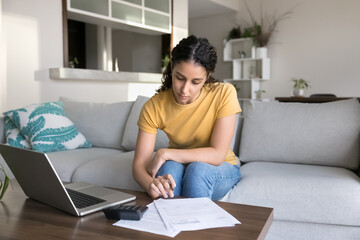 Serious Latin woman sit on couch using calculator for expenses calculation, pay utility bills using...