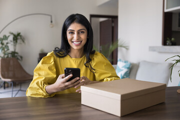 Portrait of beautiful Indian woman sit at table with parcel, using smartphone to leave positive...