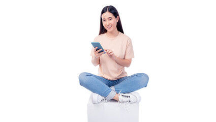 Cheerful young Asian woman sitting cross-legged on a white cube, engaging with smartphone. She wears T-shirt and blue jeans, smiling while interacting with the phone isolated on white background