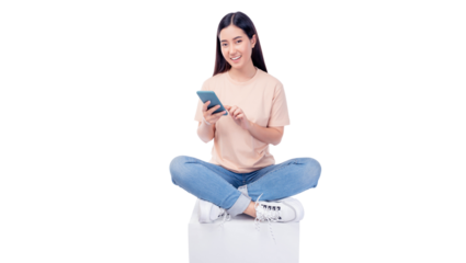 Cheerful young Asian woman sitting cross-legged on a white cube, engaging with smartphone. She wears T-shirt and blue jeans, smiling while interacting with the phone isolated on white background