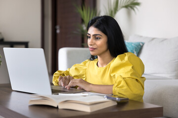 Focused Indian woman sits at table in cozy living room, studying, researching or doing professional...