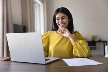 Smiling Indian woman sitting at desk with laptop, engaged in tasks completion successfully or e-learning process. Professional success, productivity, digital communication, technology-driven lifestyle