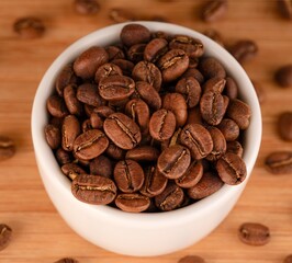 Coffee beans in a small white bowl on a wooden background near other coffee beans. Close-up
