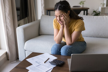 Brazilian woman sits at table with papers and laptop, feels stressed due to unpaid expenses, budgeting concerns, looks overwhelmed by debt, loans, or huge household utility bills, unexpected charges