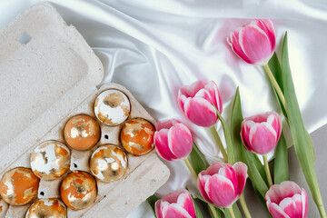 Gold Easter eggs in a carton box with pink tulips, top view. Easter festive background.