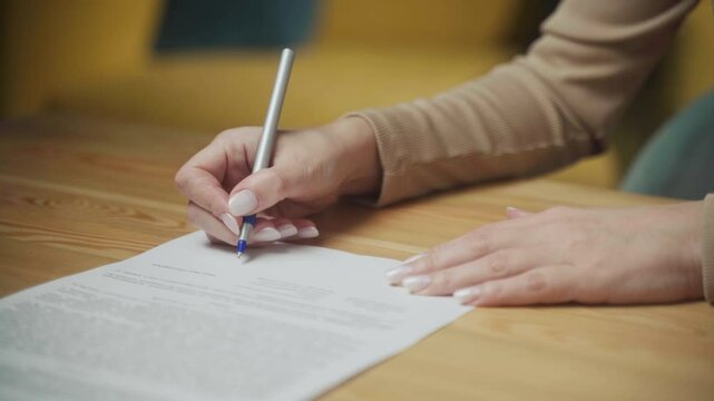 A close-up captures hands carefully signing significant documents on a wooden table. Soft natural light fills the cozy space, adding warmth to the focused activity.
