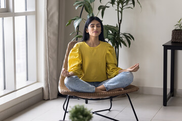 Calm Indian woman meditating at home seated in cozy armchair, practice encourages relaxation,...