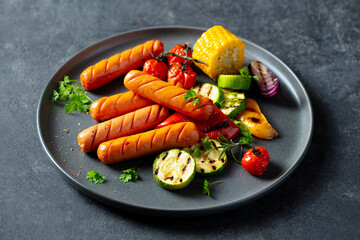 Roasted sausages with grilled vegetables, cherry tomatoes, corn and fresh herbs on a plate. Dark background. Close up.